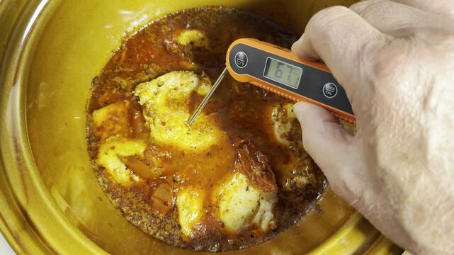 Closeup of a hand sticking the spike of a digital thermometer into a piece of chicken, which is cooking in a casserole pot, to check the temperature on the display.