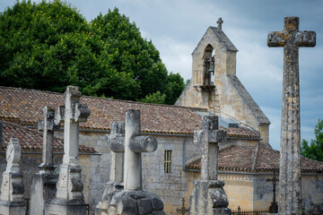 A cemetery filled with ornate stone crosses stands adjacent to a historic church in Saint-Laurent-des-Combes, surrounded by lush greenery under a dramatic sky.