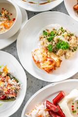 A selection of dishes on white plates, including creamy soup, baked dish with tomatoes and cheese, vegetable side with grated cheese, and sausages with mashed potatoes and pickled vegetables