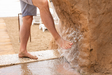 Man washing his feet with shower to clean them from the sand standing on the beach on the seashore.