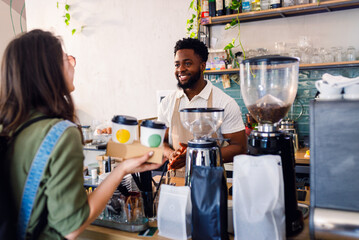 African American barista handing takeaway coffee in paper cups to female customer in cozy modern coffee shop.