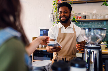 African American barista charges the customer for takeaway coffee in cozy modern coffee shop.