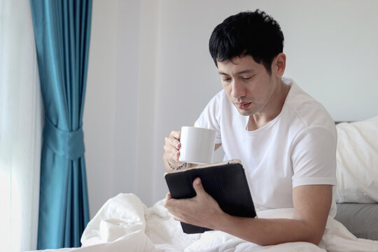 Asian man using tablet while drinking coffee in bed after waking up in morning in bedroom. Man working on his tablet during lying down on comfortable bed. Sleepy man surfing internet in bed at home.