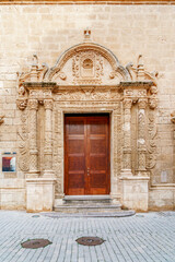 The ornate Baroque portal and grand wooden entrance door of the historic Esglesia del Roser in Ciutadella de Menorca, showcasing intricate stone carvings. This former church, now an exhibition hall