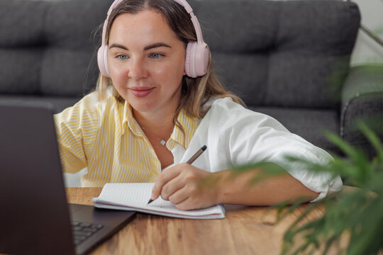 Young Caucasian woman with blond hair wearing pink headphones writing in notebook while using laptop at home desk - Powered by Adobe
