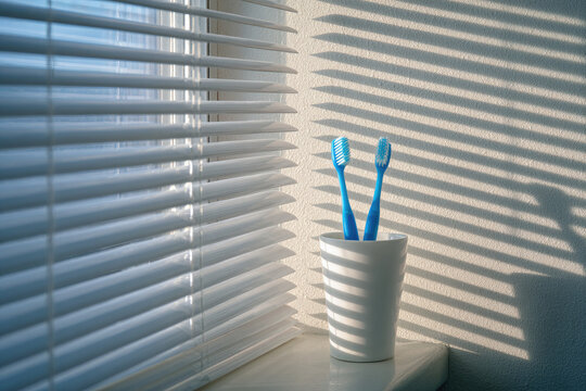 Two blue toothbrushes stand in white cup on windowsill, bathed in soft morning light filtering through blinds, casting striped shadows on wall - Powered by Adobe