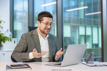 Fototapeta premium Smiling young male businessman in a suit sits at his workplace in the office and talks remotely via laptop, gesturing with his hands