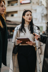 A stylish woman dressed in a cream coat holds a digital tablet while engaging in discussion with others in an urban outdoor setting, exemplifying professionalism and communication.