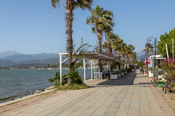 Empty tiled promenade with palm trees and lamps in Fethiye. Bay with restaurant verandas and distant misty mountains under midday sun. Fethiye, Turkey. May.