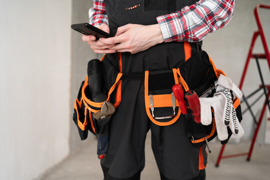 Close-up of male builder with smartphone and working tools on belt at new home background. repair, construction and building