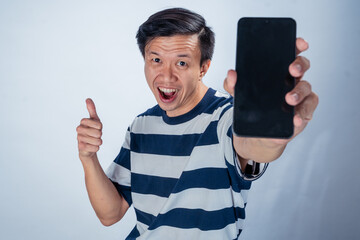 Smiling Asian man in a striped shirt holding a smartphone with a blank screen in one hand and...