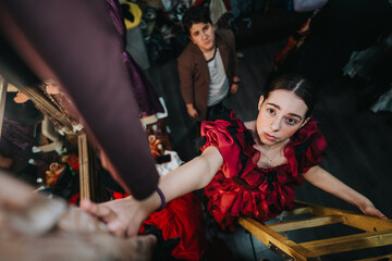 A young actress in a red costume ascends a ladder, captured from above, with a fellow actor in the background inside a theater setting.