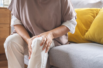 Obraz premium Body arthritis muscles stiff problem, aged woman suffering on leg, pain knee from disease chronic, hand massage rubbing joint injury, mature hurt or sore sitting on sofa at home. Health care people.