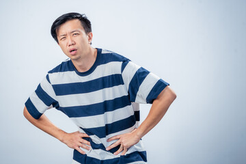 Asian man in striped shirt showing discomfort and fatigue, leaning forward with hands on waist and pained expression, standing against plain studio background