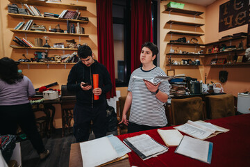 Several young individuals in a backstage area surrounded by books and props, organizing materials. The image depicts a preparation scene with engaged participants and a creative environment.