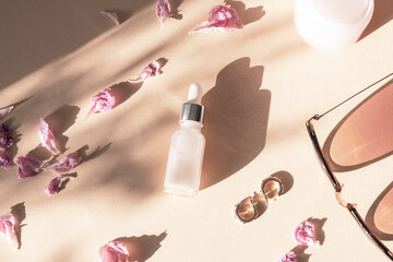 Face serum and women's accessories with dry peony petals and long hard shadows. Top view, flat lay