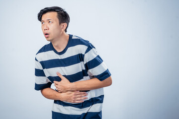 Asian man in striped shirt showing pain and discomfort while holding his chest and stomach, possibly indicating heartburn or illness, standing against plain studio background
