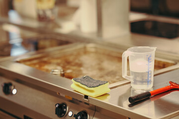 Close-up of a commercial kitchen surface with sponge, measuring cup, and cleaning scraper, illustrating cleanliness and hygiene in professional food preparation area.