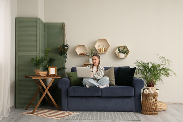 Young woman sitting on blue comfortable sofa and working with laptop in living room