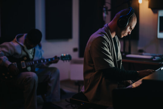 Two talented musicians collaborating in a dimly lit recording studio. One plays piano while the other plays guitar, immersed in the creative process, conveying focus and collaboration.