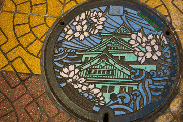 A beautifully decorated manhole cover in Osaka, Japan. It depicts cherry blossoms, a castle, and stylized waves. Vibrant colors and intricate design.