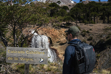 Hiker with a backpack stands near a sign for Cascada Caballera de la Virgen waterfall. Caviahue, Neuquen, Argentina