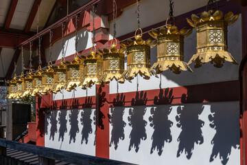 Ornate gold lanterns hang from chains, casting intricate shadows on a red and white temple wall in bright sunlight at Shitenno-ji Temple Osaka, Japan