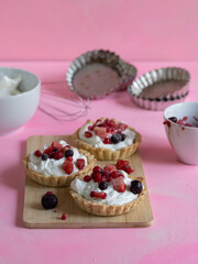 Handmade yogurt cream and red fruit tartlets on a light wooden board and with ingredients and kitchen utensils. Pastel pink background