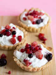 Three refreshing and healthy yogurt cream and red fruit tartlets on a light wooden board and pastel pink background