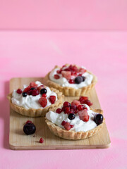 Three refreshing and healthy yogurt cream and red fruit tartlets on a light wooden board and pastel pink background