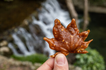 A hand holds a uniquely shaped, brown deep fried maple leaf tempura in Minoh National Park, Osaka, Japan. The blurred background shows a waterfall, hinting at a natural setting.