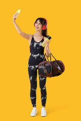 Young woman in headphones with mobile phone and sports bag taking selfie on yellow background