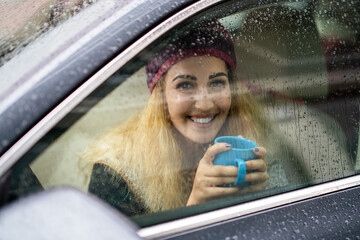 Smiling young blond woman holding coffee in the car on rainy day.