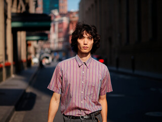 Fototapeta premium Portrait of handsome Chinese young man wearing stripe shirt posing in the street, young guy with black curly hair with urban background. Male fashion, cool Asian young man lifestyle.