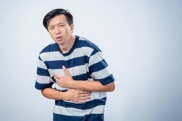 Asian man in striped shirt showing pain and discomfort while holding his chest and stomach, possibly indicating heartburn or illness, standing against plain studio background