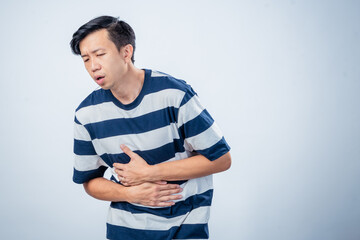 Asian man in striped shirt showing pain and discomfort while holding his chest and stomach, possibly indicating heartburn or illness, standing against plain studio background