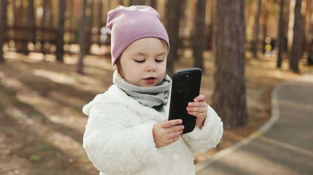 Cute child holds smartphone, looks and taps on screen, takes photo and smiles. in park at autumn time. Child takes pictures of nature. Little blogger