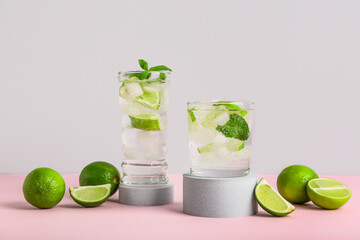 Decorative podiums with glasses of fresh Mojito cocktail and limes on pink table against grey background