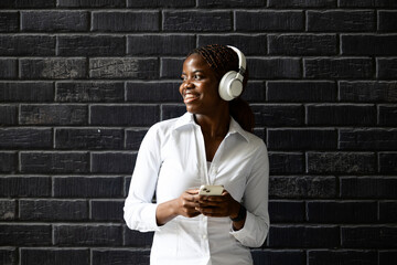 Portrait of smiling African business woman with smart phone standing by brick wall and looking sideways.