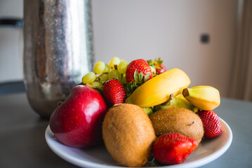 A plate of fresh fruit featuring bananas, kiwis, strawberries, grapes, and a red apple on a table. A metallic vase and blurred wall are in the background.
