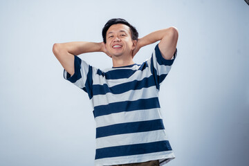 Asian man in a striped shirt smiling confidently with both hands placed behind his head, standing...