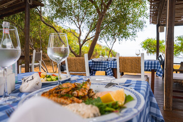A plate of grilled seafood with lemon and greens on a blue and white tablecloth, surrounded by trees, rustic decor, and a glimpse of the sea in Cyprus.