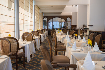 A refined dining space with neatly set tables, yellow flower centerpieces, woven back chairs, arched mirrors, and soft natural light through large windows.