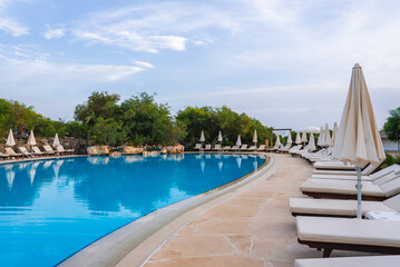 A large curved pool with clear blue water surrounded by lush greenery. White sun loungers and umbrellas line the stone paved poolside in a tranquil setting.