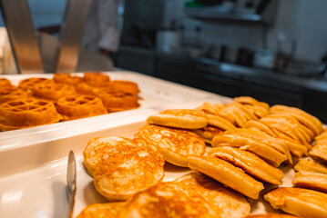 Golden brown waffles and pancakes are arranged on white trays in a buffet setup. A kitchen with stainless steel equipment is visible in the background.