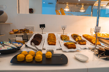 A variety of desserts, including lemon muffins and chocolate cakes, arranged on white trays in a well lit dining area with warm lighting fixtures.