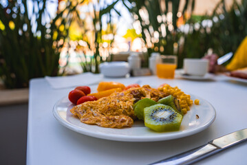 A vibrant breakfast plate with an omelet, kiwi, cherry tomatoes, and corn on a white table. Blurred greenery and a poolside setting in a sunny location.