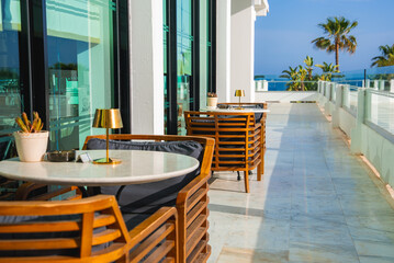 Outdoor terrace with modern wooden chairs, marble tables, gold lamps, glass railings, palm trees, and a clear view of the blue ocean in Cyprus.