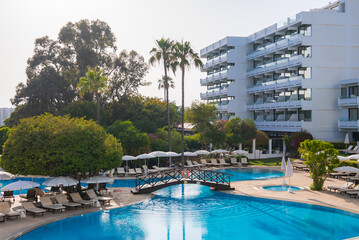 Modern white resort building with balconies, a large pool, sun loungers, umbrellas, a wooden bridge, lush greenery, and tall palm trees in Cyprus.