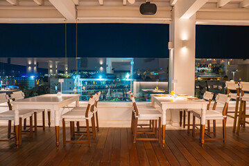Stylish indoor dining area with white wooden furniture, warm lighting, polished floors, large glass windows, and a cityscape view at night in Cyprus.